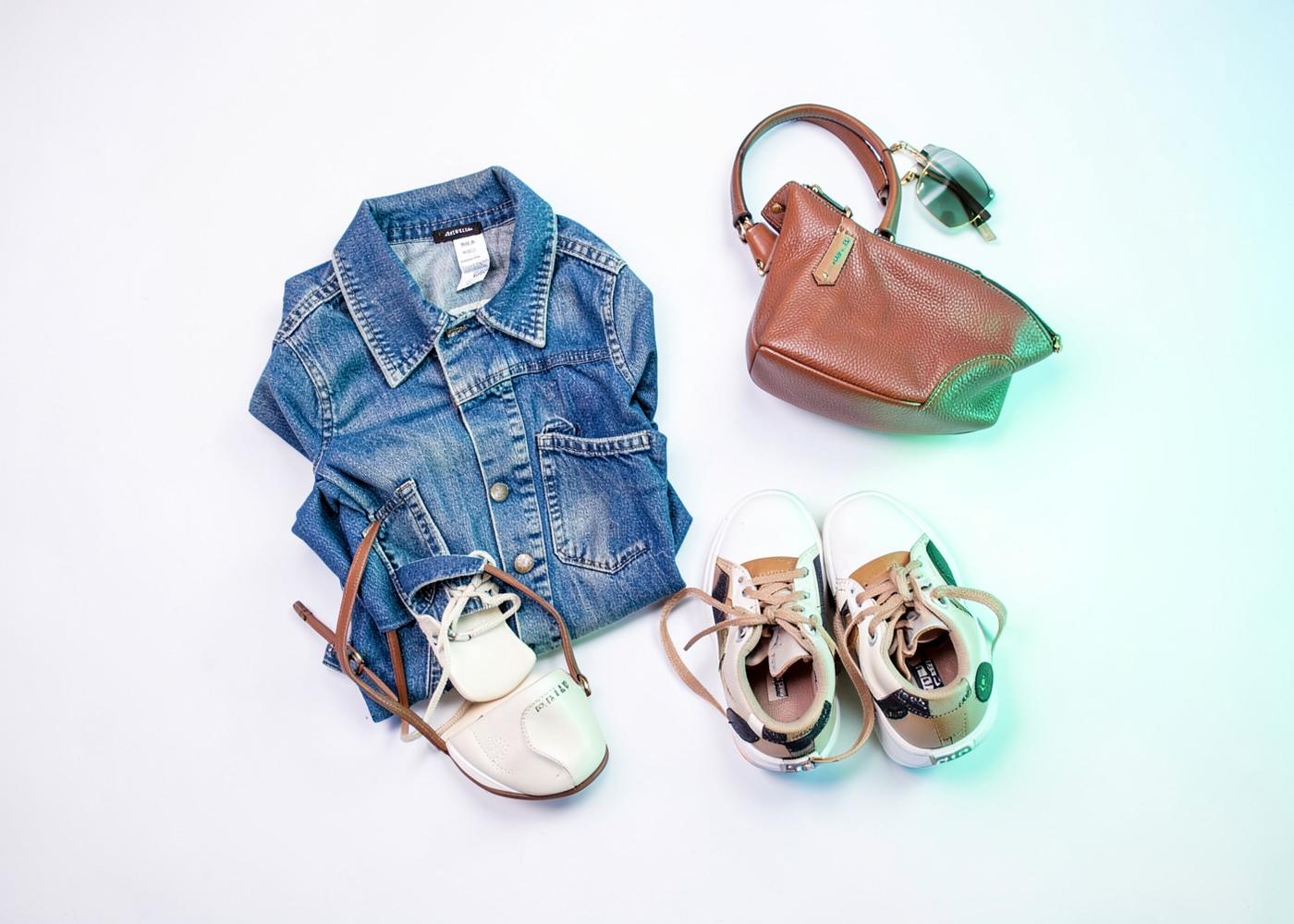 Secondhand denim jacket, leather bag, sunglasses and sneakers laid out on a white surface